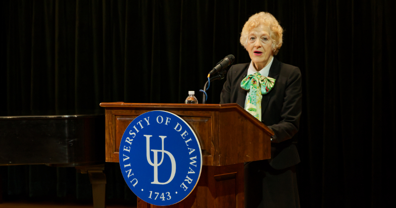 Mary Beth (M.B.) Kirkham standing behind a UD podium on stage in the Gore Recital Hall
