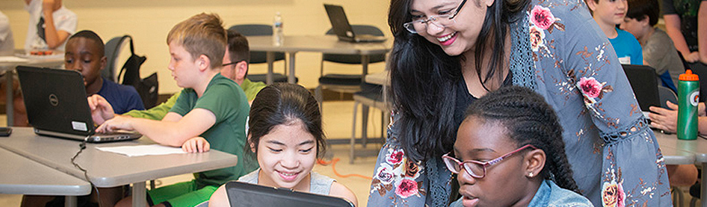 Teacher helping two elementary school students who are working together on a laptop.