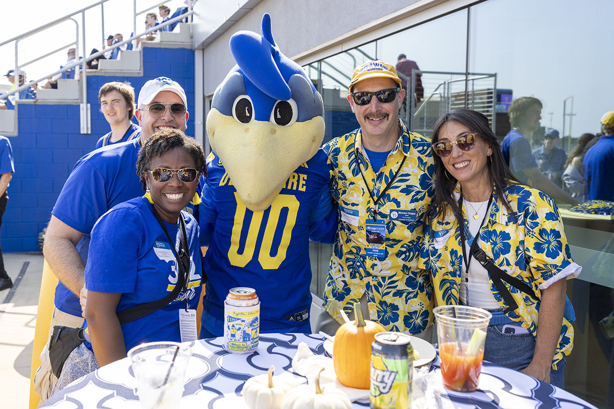 Four people. posing with the University of Delaware mascot YouDee at a football tailgate party