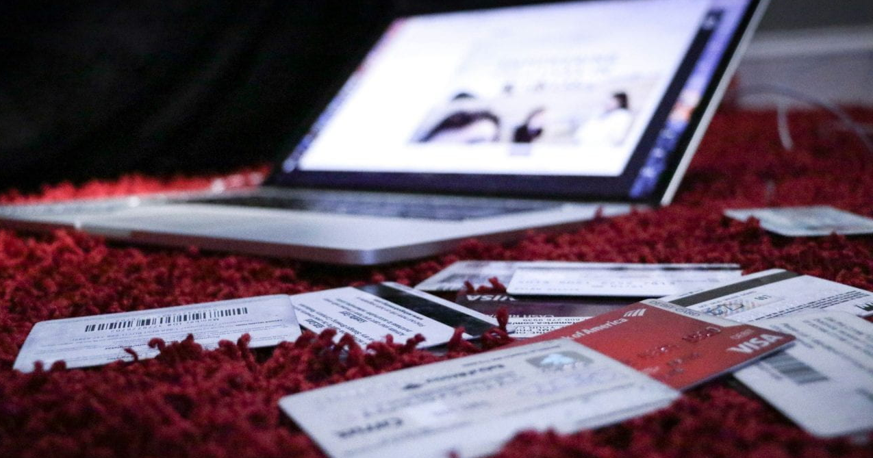 A laptop sits open on a red carpet with several credit cards and documents scattered in front of it, suggesting online shopping or financial activity. The laptop screen displays a blurred webpage with indistinct text and images.