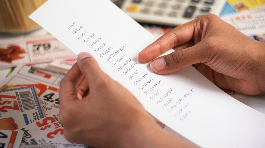 A person holding a grocery list with coupons on the table in the background.