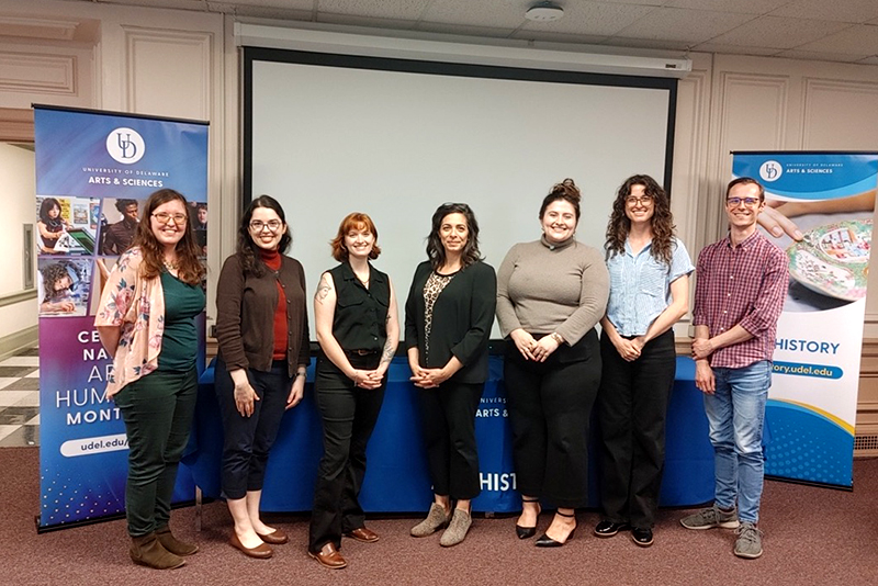 A group of seven art history graduate students standing between department signage inside a conference room with a projection screen.