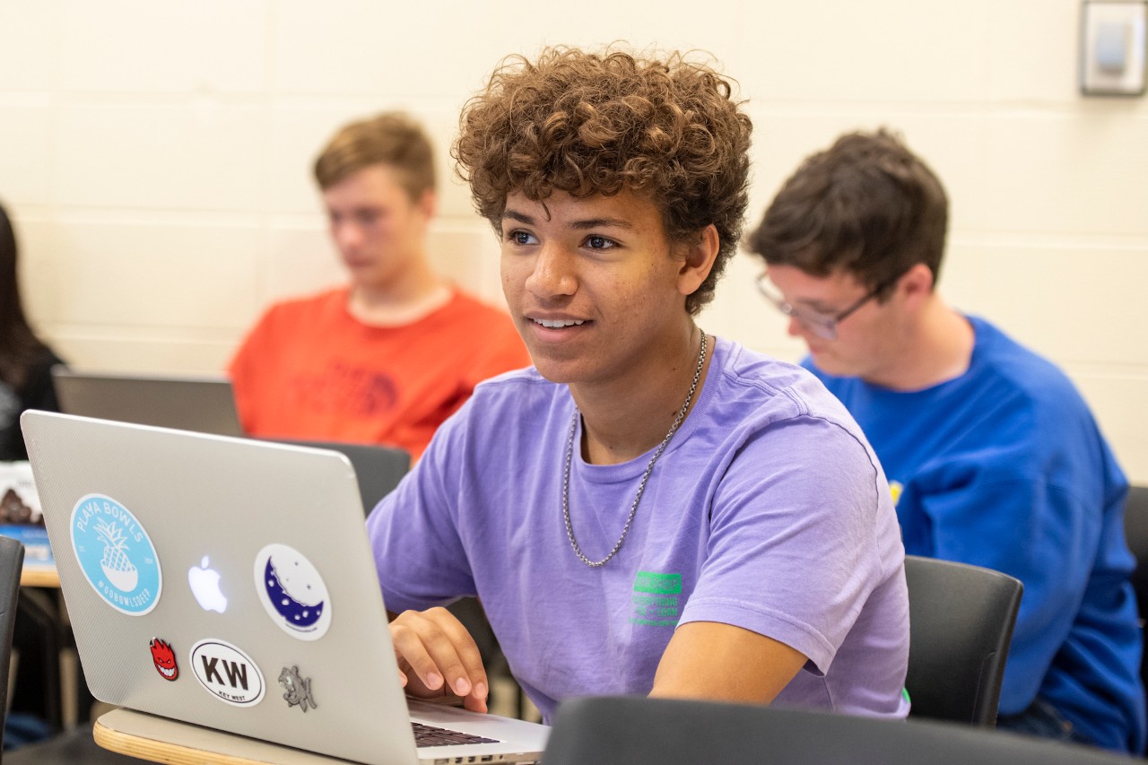 Smiling student behind a laptop. There are students in the background.