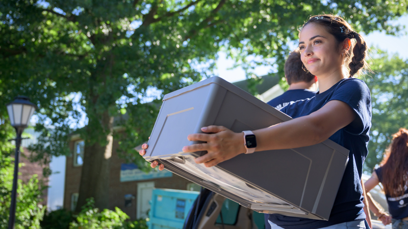 a student carries a storage bin into a residence hall on move-in day