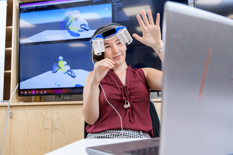 Summer_Research-COE-Christenbury-AR-VR-072318 Alina Christenbury is a sophomore working on a summer research project with assistant professor of Electrical & Computer Engineering Andy Novocin on augmented reality (AR) / virtual reality (VR) project to understand aspects of human interaction with AR / VR systems. Their test platform makes use of the Leap motion controller, a headset adapter from a welder's face shield with 3D-printed screen holders, connected to a computer running the Unity game engine. - (Evan Krape / University of Delaware)
