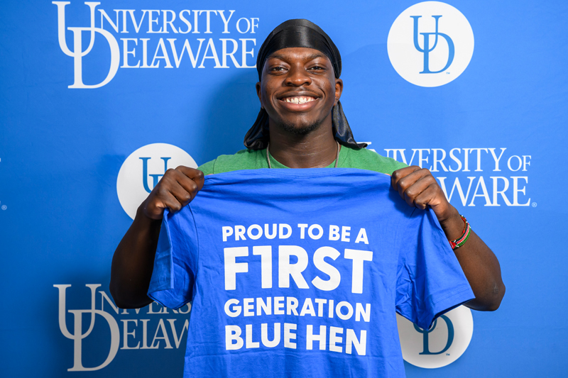 a smiling student holds up a t-shirt reading "PROUD TO BE A F1RST GENERATION BLUE HEN"