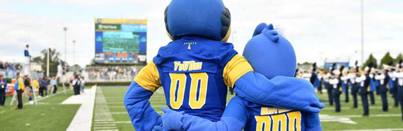 YouDee and Baby Blue stand on the sidelines with their arms around each other watching the marching band perform.