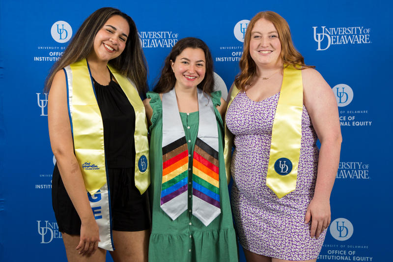 three students wearing first-generation graduation stoles pose in front of a blue UD background