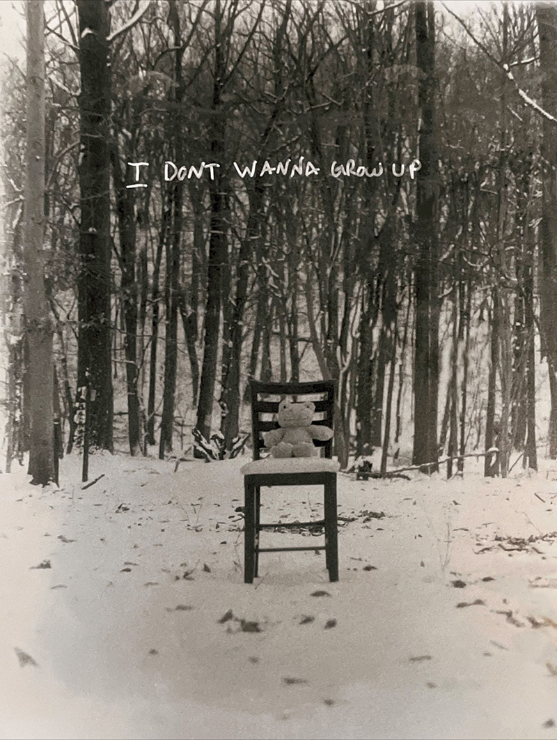 Black and white photograph of a teddy bear on a chair in front of a snowy forest of bare trees. The words "I don't wanna grow up" are superimposed on the photo.