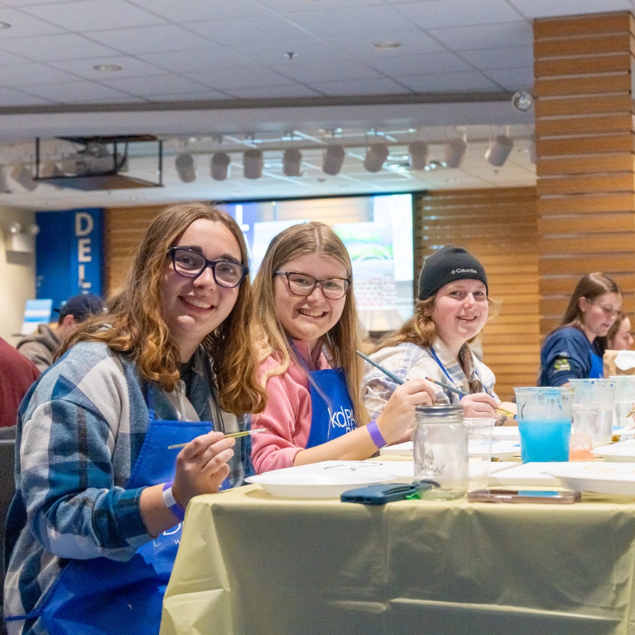 students smile at a table while painting on canvases