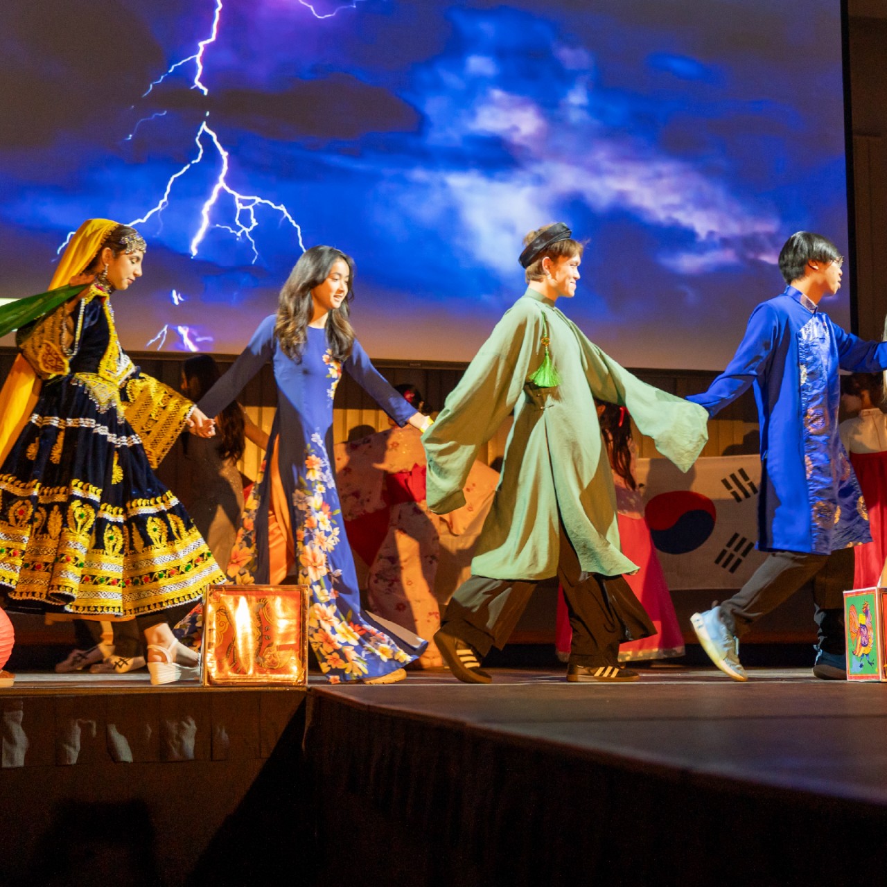students in Asian attire cross a stage holding hands.