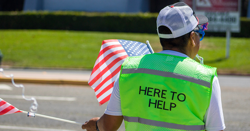 Man holds American flag wearing neon vest that reads "here to help"
