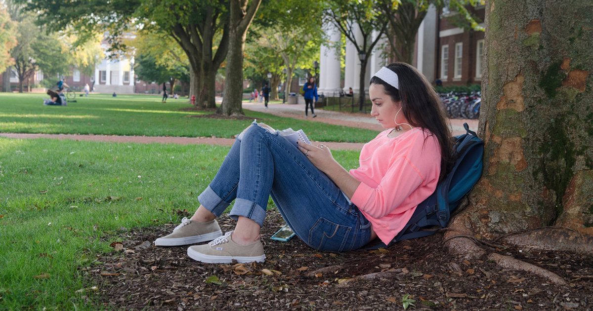 Female student sits reading against a tree on The Green