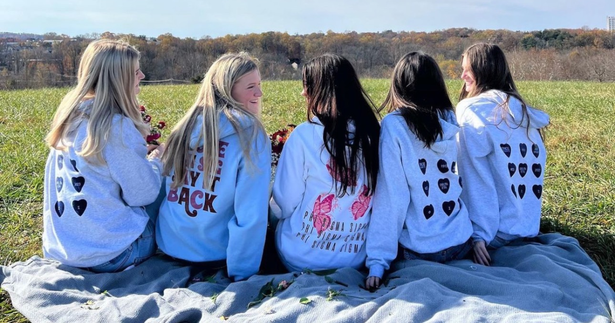Five Phi Sigma Sigma sorority sisters sit on blanket on grassy hill overlooking trees