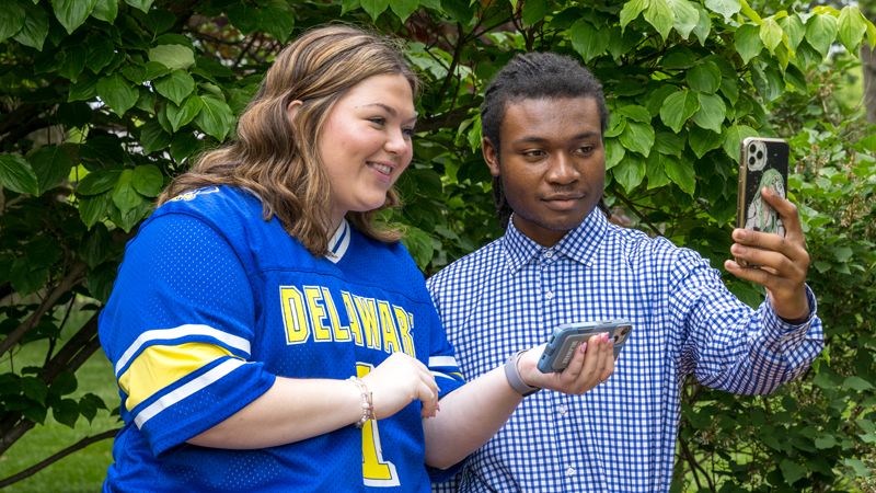 two students stand outside and smile while viewing information on their phones