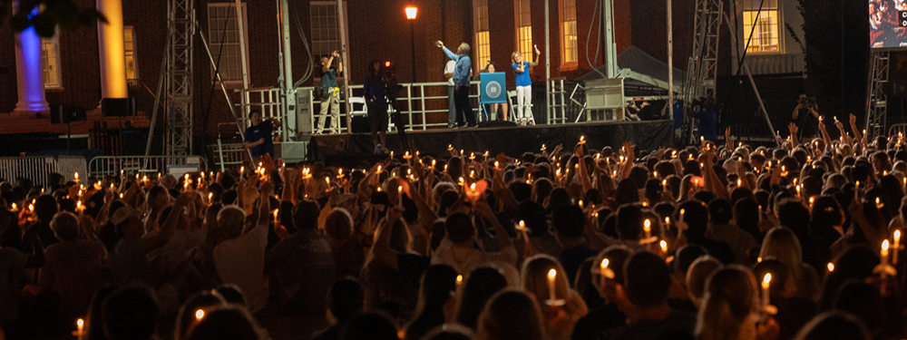 a wide view of students holding lit candles toward President Assanis on the stage at the Welcome Days Twilight Induction Ceremony