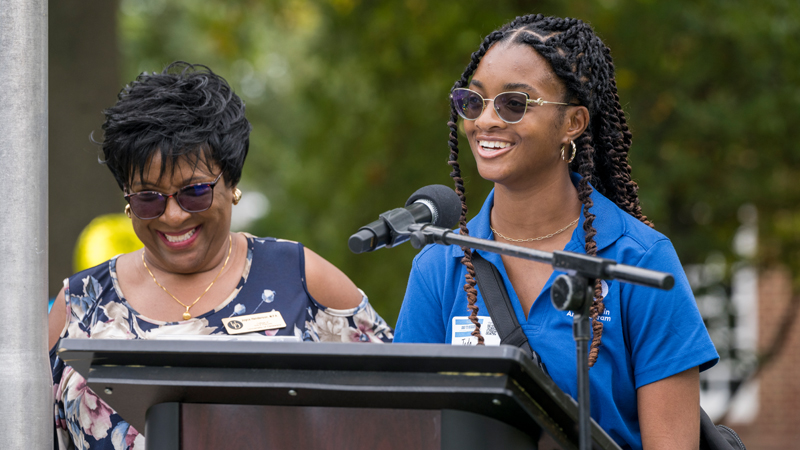 a smiling student stands at a podium speaking to an audience while a staff member stands behind her.