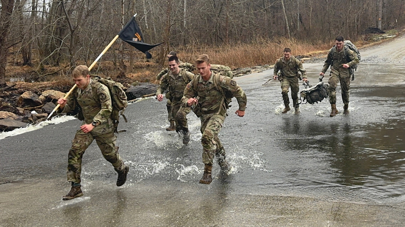 cadets in the AROTC run through water on a trail; one is carrying a flag.