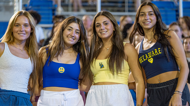 four female students pose in UD gear at a UD football game.