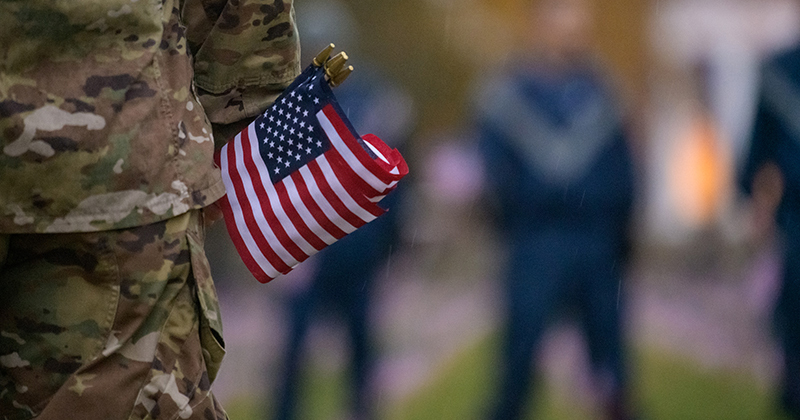 a student in camouflage military uniform stands with small American flags in their hand