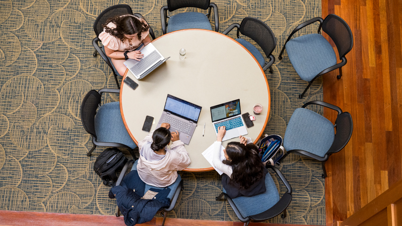 an overhead view of three students sitting at a round table, each working on a laptop.