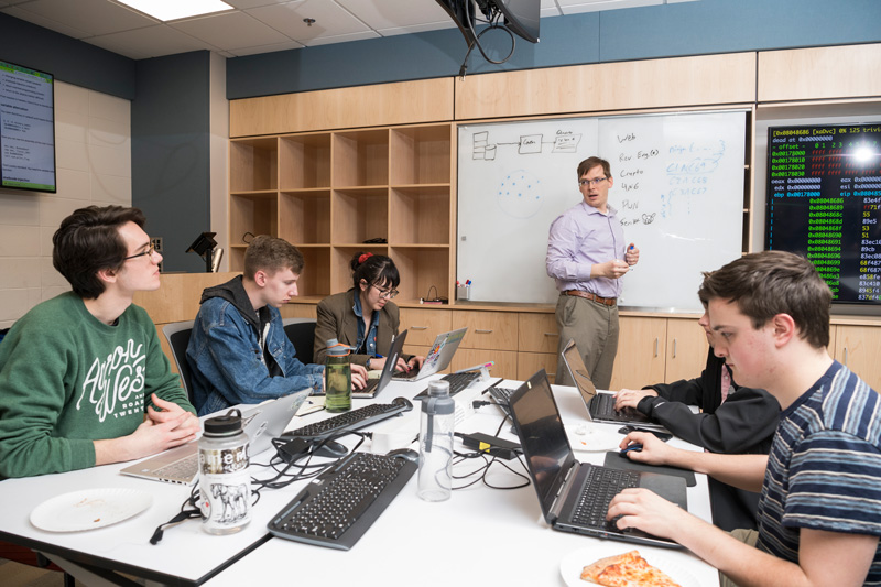 Dr. Andy Novocin teaches students developing and practicing cybersecurity skills to make them more “ethical” hackers in the world of cybersecurity in the iSuite at Evans Hall, Friday, March 22nd, 2019.  Students are: Dan Goodman (gray long sleeve), Vineeth Gutta (black shirt, khakis), Landon JOnes (black jacket), Ryan Geary (green crewneck), Thomas Pisklak (striped t-shirt), Isabel Navarro (red pants, brown blazer), Collin Clark (jean jacket, nose piercing), And Alena Gusakov (red shirt, jeans)  (PHOTO RELEASES WERE OBTAINED ON ALL PICTURED.)