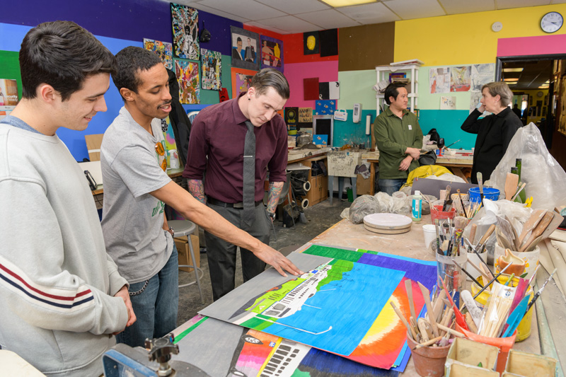 Joshua Stout, a doctoral student in sociology (maroon shirt w/ tie) and Dillon DiGuglielmo, a senior criminal justice major (light-gray sweater) looking over artist Geraldo Gonzalez's (gray short-sleeve tee) work at the Creative Vision Factory (CVF). Stout and DiGuglielmo worked on a project to create a digital archive of CVF members’ work (including Gonzalez's). The CVF is downtown Wilmington community arts organization providing work and exhibition space to individuals on the behavioral health spectrum  - (Evan Krape / University of Delaware)