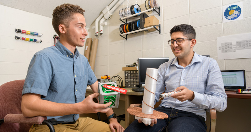 CAS-PHYS-Summer_Research-CURIE_Mission-072419 Ahmad Jalal Al Tamini (long sleeve shirt) and Jeffrey Neumann (short sleeve shirt) are undergraduate students working on a Summer Research project to help implement an interface to receive data from the “Cubesat Radio Interferometry Experiment (CURIE).” Lead by researcher Edward Graff (Physics) with Faculty Mentor Bennett Maruca, (Physics and Astronomy) the project aims to develop and deploy a ground station to up- and downlink data with the two CURIE CubeSats which will be making interferometric observations of the Sun. The ground station will be installed at the Mount Cuba Observatory in Wilmington, DE.
Pictured: Jeffrey holds a 3D printed model of a 1:1 scale CubeSat module. The CURIE mission satellies are the same width and length and only 3 times as high.
