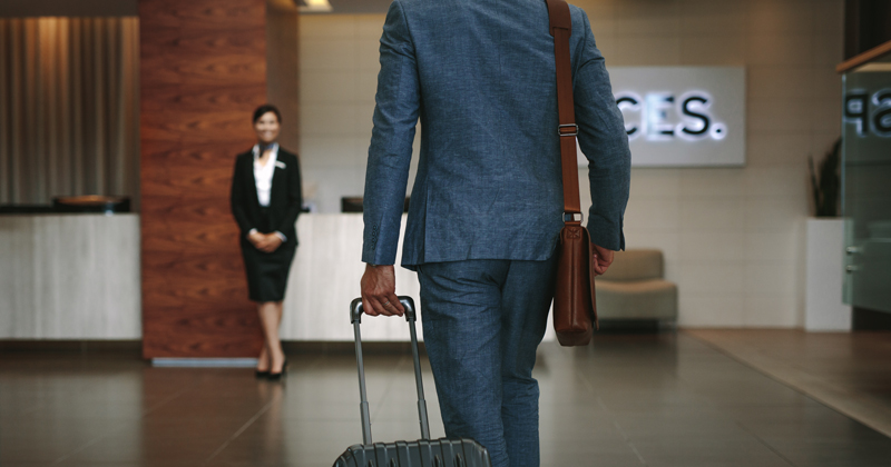 Businessman carrying suitcase while walking in hotel lobby. Business traveler arriving at hotel with female receptionist standing in background for welcoming.