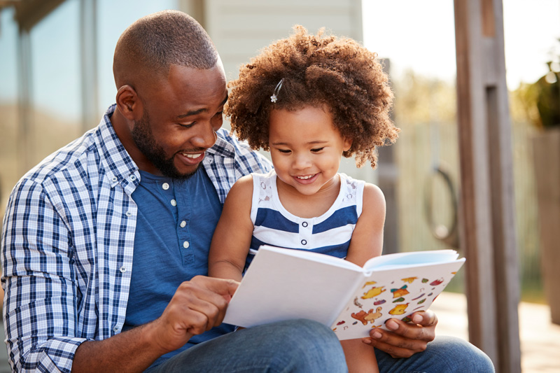 947854276 Young black father and daughter reading book outside