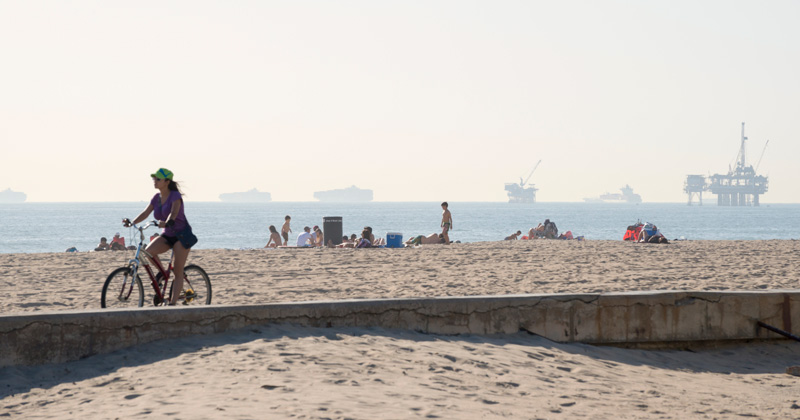 Beyond the bikeway and beach at Huntington Beach in Orange County, California, oil rigs rise from the ocean and container ships wait to enter the nearby port of Long Beach