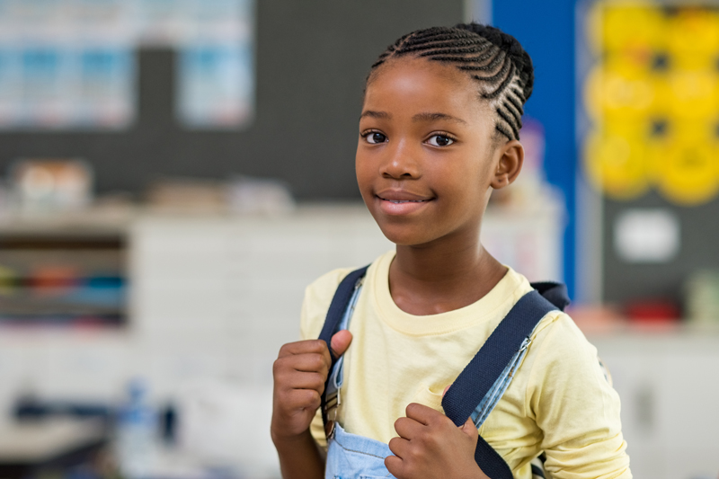 African young girl with blue backpack looking at camera. Pretty and satisfied black schoolgirl with rucksack smiling in class. Portrait of beautiful school girl standing in classroom.