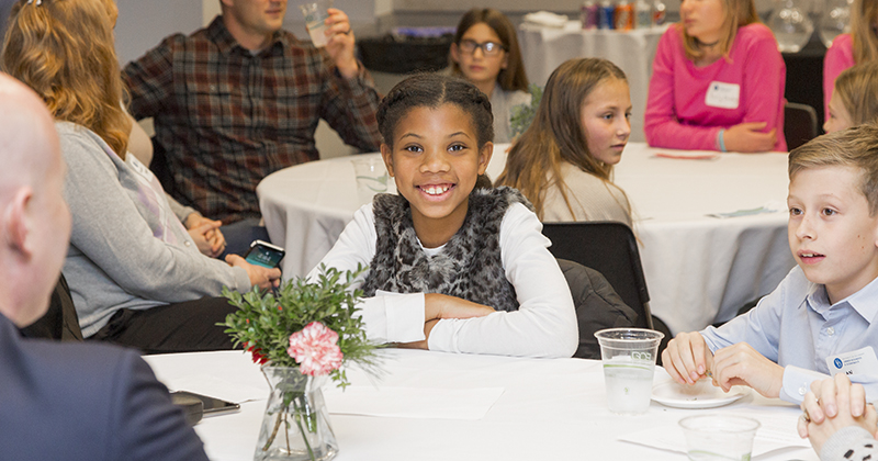 Lerner-CEEE-Stock-Market-Game-Awards-011420 Student smiles at camera while sitting at a table surrounded by fellow studnets.