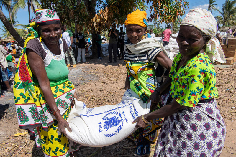 Three women carries a bag of grain as World Food Programme (WFP) distributes food in Nacate village, near Macomia.This photograph is part of a series of images made in the days leading up to the arrival of Secretary-General António Guterres in Mozambique. Mr. Guterres visited the country to take stock of the recovery efforts in the areas impacted by cyclones Idai and Kenneth, which hit just a few weeks apart in March and April 2019. More than 600 people perished during Idai alone; the effects of the two cyclones combined left approximately 2.2 million people in need of assistance. The United Nations and its humanitarian partners have since been on the ground supporting the Government’s efforts - assisting through contributing to the coordination of international support, distributing food, drinking water and medicine, and providing shelter to those displaced.