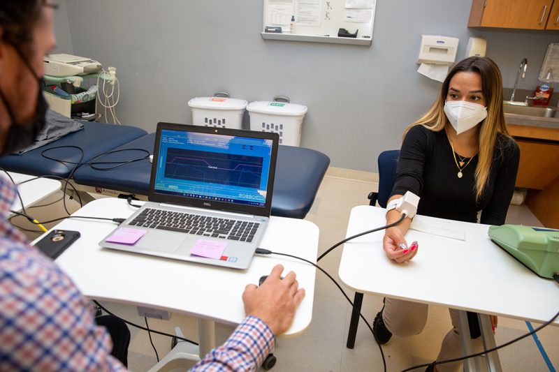 Ashley Fath, clinical research coordinator for UD’s Rehabilitation and Neuroplasticity Lab, wears a device that emits heat to induce pain as researchers seek to determine how sensitive someone might be to heat-based pain as part of a larger study on pain and motor learning. Graduate research assistant and doctoral student Patrick Knox (left) is analyzing pain levels on the screen. 