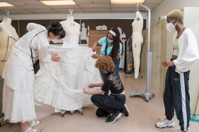 Katya Roelse (left) and Kayla Brown hold rosettes up to the gown as Alex Culley (kneeling) checks their placement on the skirt and Maya Bordrick (far right) observes the work.