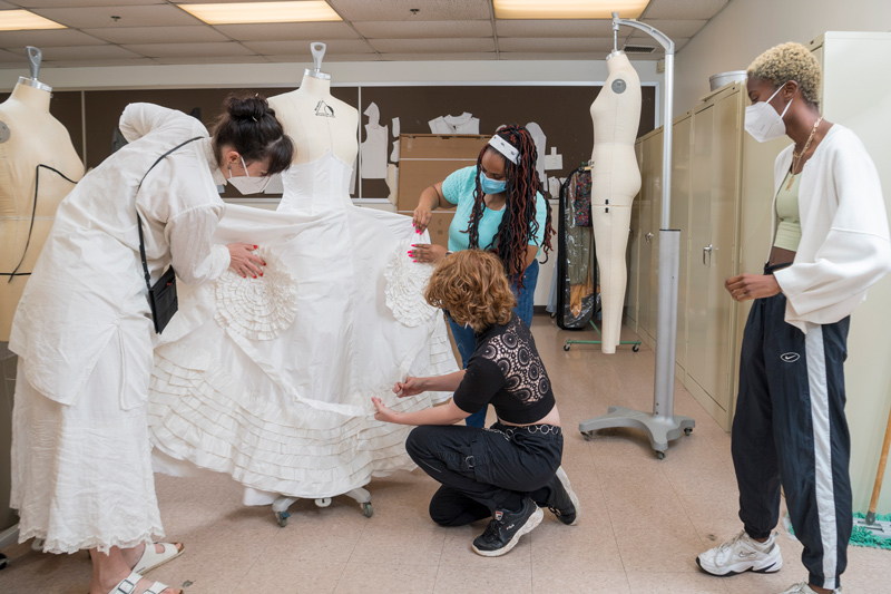 Katya Roelse (left) and Kayla Brown hold rosettes up to the gown as Alex Culley (kneeling) checks their placement on the skirt and Maya Bordrick (far right) observes the work. Katya Roelse (left) and Kayla Brown hold rosettes up to the gown as Alex Culley (kneeling) checks their placement on the skirt and Maya Bordrick (far right) observes the work.
