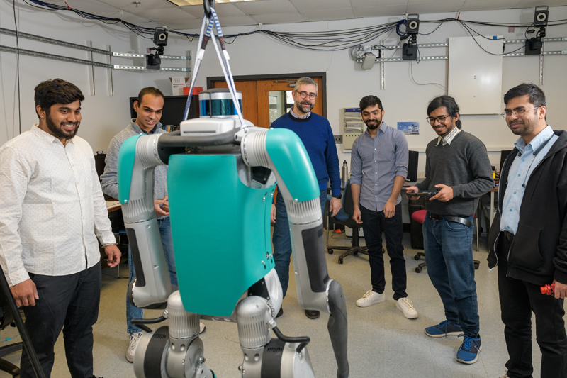 A $250,000 teal robot named Digit (shown in the forefront) will help a team of UD engineers improve two-legged robots’ ability to walk on changing terrain. Shown are (left to right) mechanical engineering graduate students Aditya Shreyas, Prem Chand, Prof. Ioannis Poulakakis, Dhruv Ashwinkumar Thanki, Abhijeet Kulkarni and Kunal Sanjay Narkhede.