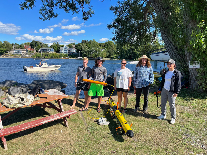 Participants in the UD AUV Bootcamp including UD graduate students and U.S. Naval Academy interns stand holding the Iver3 AUV magnetometer while the R/V Parker passes behind at a Sackets Harbor N.Y. site of conflict during the War of 1812.