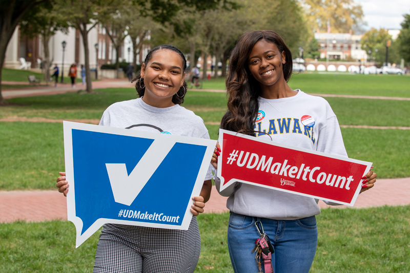 Two students holding signs encouraging students to register to vote. Beginning Tuesday, Sept. 13, UD student groups and organizations will compete to register new voters through TurboVote, a web application that helps eligible U.S. citizens register to vote, request an absentee ballot, and sign up for Election Day text and/or email reminders.
