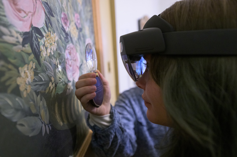 Margalit Schindler holds a magnifier up to a painting so that Morrigan Kelley, wearing the augmented reality device, can transmit details for Joelle Wickens to see on her laptop.