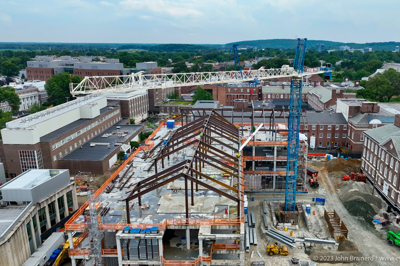A drone’s eye view of Building X (looking south from East Delaware Avenue) shows the last piece of structural steel nearing its final destination. It is the slender white beam at the center of the image. Two workers in bright yellow shirts are waiting to secure it.