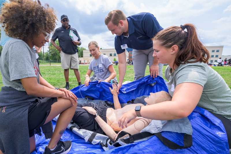 Delaware high school athletic trainers (AT) take part in a hands-on professional development workshop led by UD alumnus Darryl Conway (standing, back center) ahead of the fall sports season. In this drill, athletic trainers are faced with a scenario involving a cross-country runner who passed out from exertional heat stroke. They work to cool the athlete as quickly as possible via a whole-body ice-water immersion technique.