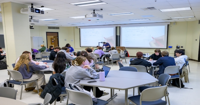 CTAL-Open_Teaching_Day-MATH231-040225 Students sitting at octagonal tables in a classroom with two projector screens featuring calculus problems.