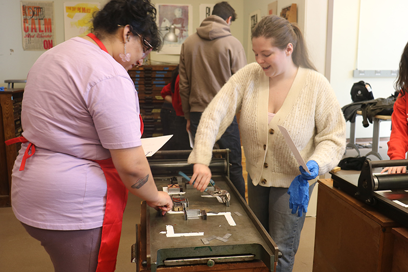 Spring 2025 art graduates Lillyanne Dominguez and Cassidy Driscoll use magnets to keep tiles in place on one of Raven Press’s hand press machines, trying to achieve the “perfect kiss.” 