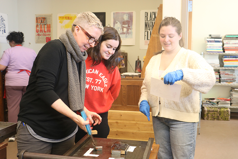 Associate Professor of English Laura Helton shows Gia Joella and Cassidy Driscoll how to place tiles on the hand press. If the tiles move when the roller is pulled across, the printed image and letters will smear. 