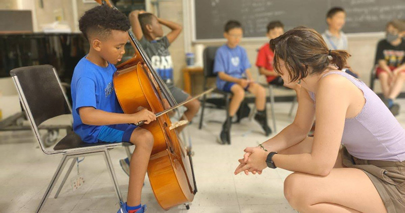CMS Administrative Assistant and teacher Emme Whisner introduces a young student to the cello during Meet the Instruments camp. 