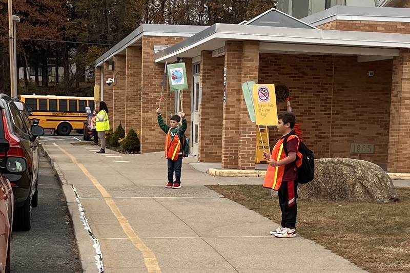 Elementary school students encourage parents and caregivers to turn off their car engines while waiting for their children in the school’s carline. 