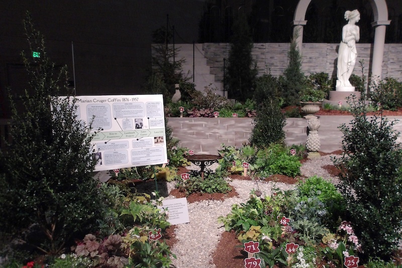 A birdbath is in the center of this photo. Some interesting plants featured here are the bergenia flowers — pink flowers in the bottom right, the hellebores (No. 71) and the red veiny foliage of the heuchera (bottom left of the photo). 