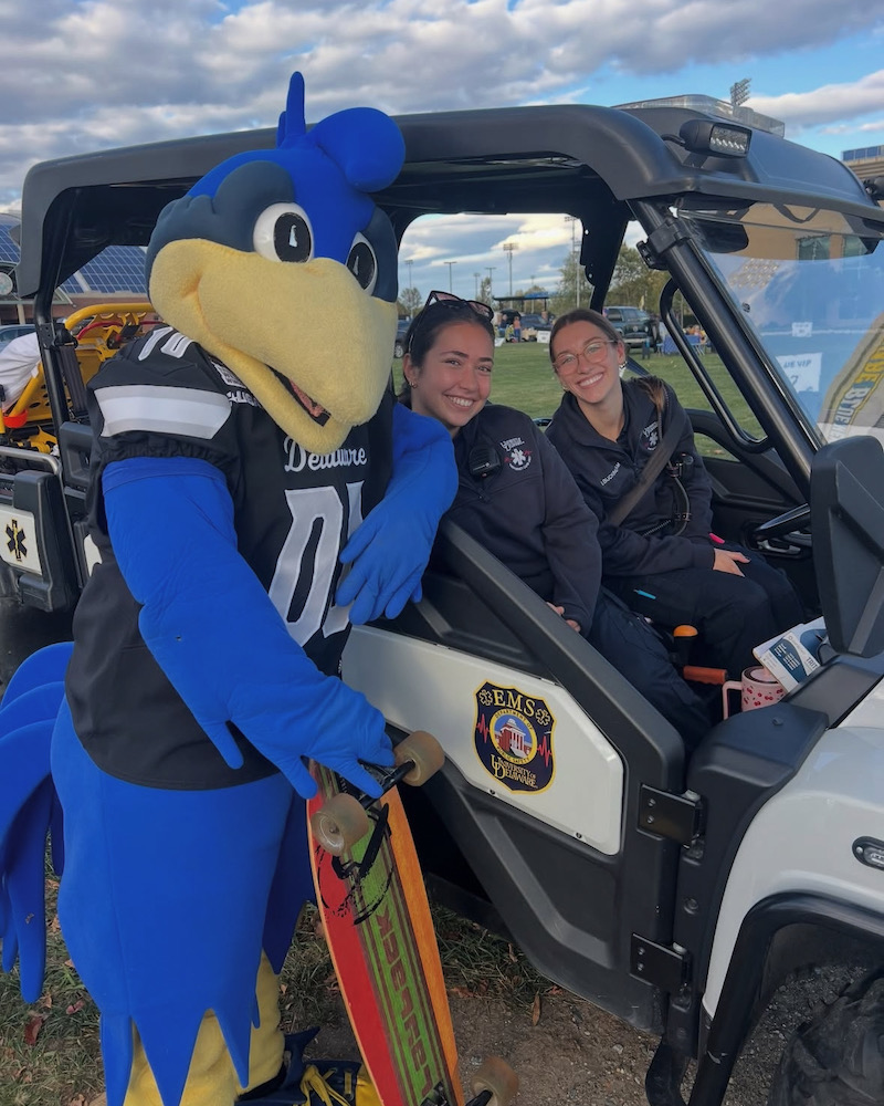 YouDee visits with UD students — and volunteers of student-run UDECU — Tamar Peleg (center) and Sarah Buckingham at a 2025 UD football game. At each game, UDECU oversees an EMS command post, and staffs a full ambulance crew, an EMS Gator and bike teams to ensure safety and swift response.